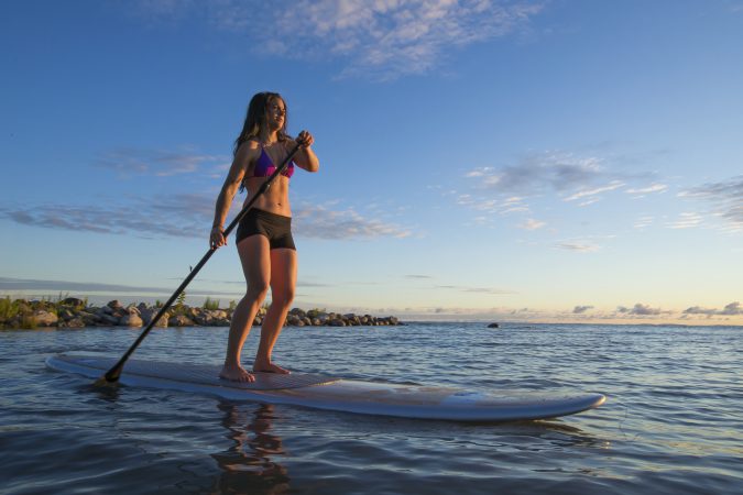 A lone woman paddles her stand up paddle board through the waves as the sun rises.